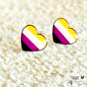 May include: A pair of heart-shaped earrings with a non-binary pride flag design. The earrings feature yellow, white, purple, and black stripes. The earrings are set against a textured, off-white background.