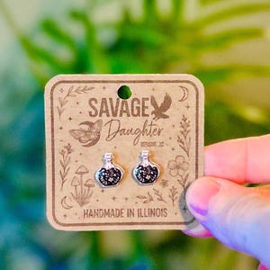 May include: A pair of silver-toned stud earrings shaped like small potion bottles with black and white floral details. The earrings are attached to a square brown cardboard display card featuring the text Savage Daughter Designs LLC.