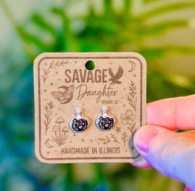 May include: A pair of silver-toned stud earrings shaped like small potion bottles with black and white floral details. The earrings are attached to a square brown cardboard display card featuring the text Savage Daughter Designs LLC.