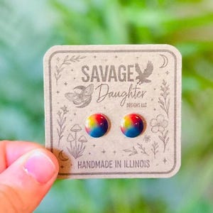 May include: A pair of rainbow-colored stud earrings on a decorative card. The card features the text "SAVAGE DAUGHTER DESIGNS LLC" and "HANDMADE IN ILLINOIS" with botanical illustrations. The earrings have a glossy finish.