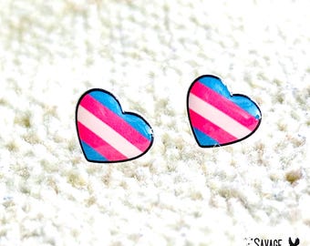 May include: Two heart-shaped earrings with a transgender pride flag design. Each earring features a blue top and bottom, pink center stripes, and white stripes. The earrings have a black outline and are set against a white, textured background.
