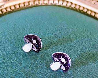 May include: A pair of stud earrings shaped like mushrooms. The mushroom caps are black with a white triple moon symbol and small white dots representing stars. The stems are solid white. The earrings have a flat, graphic design style.