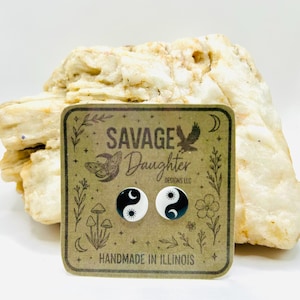 May include: A pair of black and white yin yang stud earrings on a brown card with the text "SAVAGE DAUGHTER DESIGNS LLC" and "HANDMADE IN ILLINOIS". The earrings are displayed in front of a light-colored, textured rock.