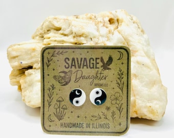May include: A pair of black and white yin yang stud earrings on a brown card with the text "SAVAGE DAUGHTER DESIGNS LLC" and "HANDMADE IN ILLINOIS". The earrings are displayed in front of a light-colored, textured rock.