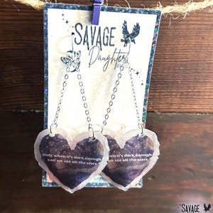 May include: Heart-shaped earrings with a celestial design and the text "Only when it's dark enough can we see all the stars." The earrings are suspended from a chain and attached to a decorative card with the text "SAVAGE Daughter".