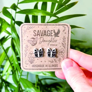 May include: A pair of black and white gift box earrings with a bow design. The earrings are on a brown card with the text "SAVAGE Daughter DESIGNS LLC" and "HANDMADE IN ILLINOIS". The card has illustrations of a butterfly, mushrooms, and flowers.