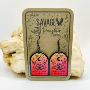 May include: A pair of black metal earrings with a celestial design. Each earring features a moth, crescent moon, and sun design in shades of red, orange, and yellow. The earrings are displayed on a brown card with the text "SAVAGE Daughter Designs".