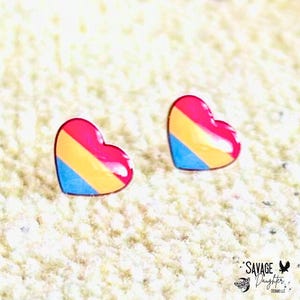 May include: Two heart-shaped earrings with a glossy finish. Each earring features a design with pink, yellow, and blue stripes. The earrings are set against a textured, off-white background.