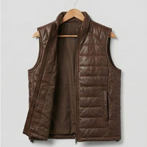 May include: Brown leather vest with a quilted pattern, a stand-up collar, and a full-length zip. The sleeveless vest has a pocket on the lower right side. It hangs on a wooden hanger against a plain background.