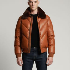 Op de afbeelding: Een cognac-kleurige leren donsjack met een chevron-gewatteerd patroon, een donkerbruine bontkraag, een ritssluiting aan de voorzijde en geribbelde gebreide boorden. Het jack wordt gedragen over een zwart shirt en een donkerblauwe spijkerbroek.