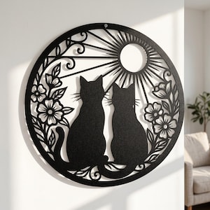 Puede incluir: Una pieza de arte mural circular de metal negro con un diseño cortado con láser de dos gatos sentados uno al lado del otro. Los gatos están rodeados de motivos florales y un patrón de rayos de sol con un sol central en un marco.