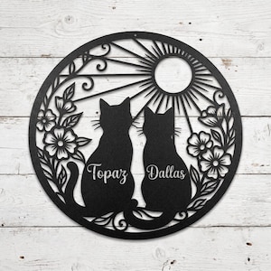 Op de afbeelding: Een cirkelvormig wandornament van zwart metaal met de silhouetten van twee katten die naast elkaar zitten. Het ontwerp bevat bloemmotieven aan de zijkanten en een zonnestraalpatroon aan de bovenkant. De namen Topaz en Dallas staan op de katten.