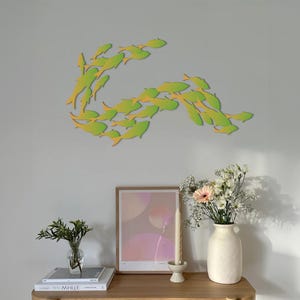May include: A wall decor featuring a school of green and yellow fish in a swirling pattern. The fish are cut out of paper and mounted on a light gray wall. The decor is displayed above a wooden shelf with a vase of flowers, a candle, and a framed print.