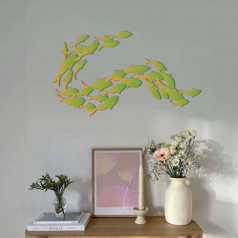 May include: A wall decor featuring a school of green and yellow fish in a swirling pattern. The fish are cut out of paper and mounted on a light gray wall. The decor is displayed above a wooden shelf with a vase of flowers, a candle, and a framed print.