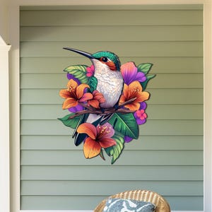 May include: A colourful hummingbird with green and white feathers perched on a branch with pink, orange and purple flowers. The hummingbird is surrounded by green leaves.