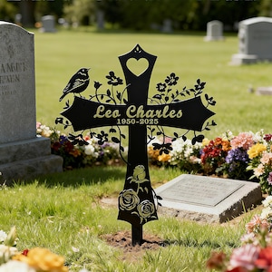 May include: A black metal cross with a heart cutout and floral detailing, set in a cemetery. The cross bears the name "Leo Charles" and dates "1950-2025". A bird is perched on the cross.