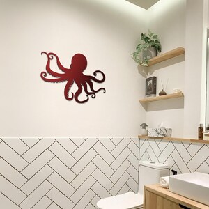 May include: A red metal octopus wall sculpture in a bathroom setting. The octopus is mounted on a white wall above a white tiled herringbone pattern. The octopus has eight tentacles and is in a curled position.