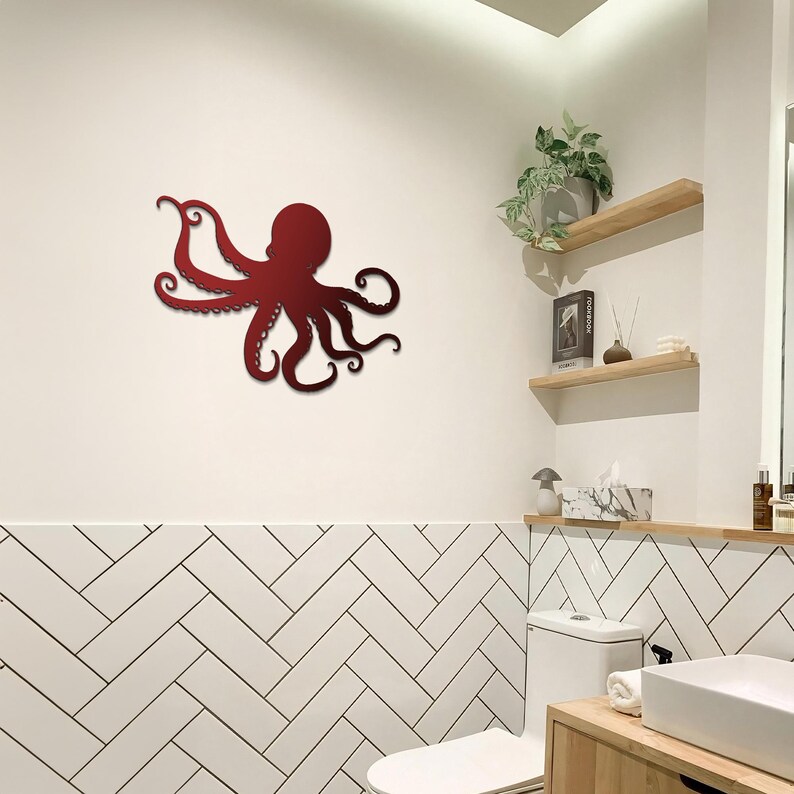 May include: A red metal octopus wall sculpture in a bathroom setting. The octopus is mounted on a white wall above a white tiled herringbone pattern. The octopus has eight tentacles and is in a curled position.