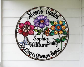 May include: A round metal garden sign with a black metal frame and colourful flowers and butterflies. The sign reads "Mom's Garden" with the names "Sophia William Emily" and the phrase "Love Grows Here".
