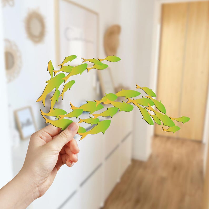 May include: A hand holds a school of green and yellow fish cutouts, arranged in a curved shape. The fish are made of paper or cardboard and are a popular craft project for children.
