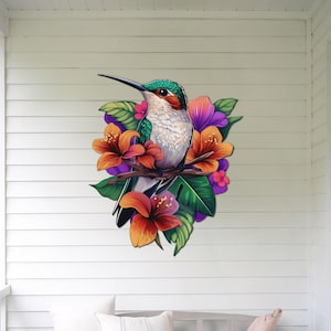 May include: A green and grey hummingbird with a long beak perched on a branch with orange, pink and purple flowers. The hummingbird is surrounded by green leaves.