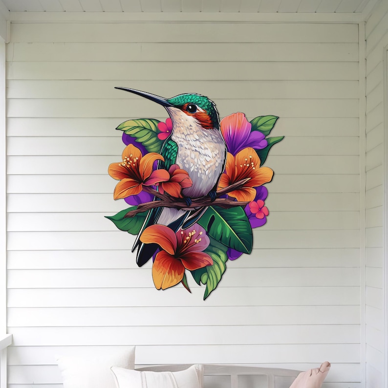 May include: A green and grey hummingbird with a long beak perched on a branch with orange, pink and purple flowers. The hummingbird is surrounded by green leaves.
