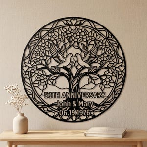 May include: A circular black wall art piece featuring a laser-cut tree design with two doves perched in the branches. The text reads 50TH ANNIVERSARY, John & Mary, and 06.19.1976. The intricate cutout pattern includes leaves and heart shapes.