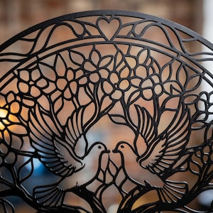 May include: A circular black metal wall art piece featuring a laser-cut design of two doves facing each other. The birds are positioned beneath a canopy of stylized flowers and branches, with a small heart shape at the top center of the frame.