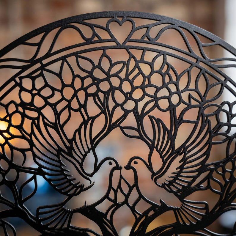 May include: A circular black metal wall art piece featuring a laser-cut design of two doves facing each other. The birds are positioned beneath a canopy of stylized flowers and branches, with a small heart shape at the top center of the frame.