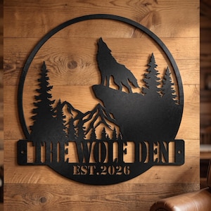 May include: A circular black metal wall sign featuring a silhouette of a howling wolf on a cliff, pine trees, and mountain peaks. The sign includes the text THE WOLF DEN and EST. 2026 cut out from the bottom section of the circular frame.