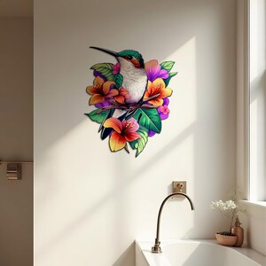 May include: A colourful hummingbird with green and white feathers perched on a branch with pink, orange and yellow flowers. The hummingbird is surrounded by green leaves.