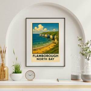 May include: Framed art print of Flamborough North Bay, featuring white cliffs, green grass, and a sandy beach. The artwork shows a blue ocean and sky. The print is in a black frame and is displayed on a white shelf, evoking a coastal theme.