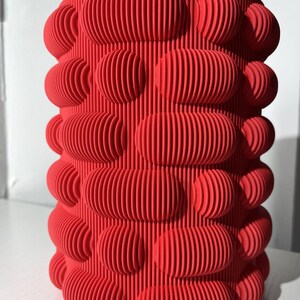 Könnte beinhalten: Ein roter zylindrischer Topf mit einer strukturierten Oberfläche. Die Oberfläche besteht aus sich wiederholenden ovalen Formen mit vertikalen Linien.
