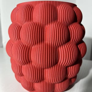 Könnte beinhalten: Ein roter, 3D-gedruckter Pflanztopf mit einem einzigartigen, wellenförmigen, strukturierten Design. Der Topf ist aus Kunststoff und hat eine runde Form.