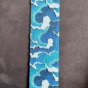 Könnte beinhalten: Ein rechteckiges Lesezeichen mit einem geschichteten, dreidimensionalen Design aus stilisierten Meereswellen. Das Kunstwerk verwendet eine Palette aus tiefem Marineblau, Königsblau, hellem Türkis und Weiß mit feinen Wellenlinien.
