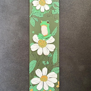 Könnte beinhalten: Ein grüner Lesezeichen mit einer Frosch-Illustration. Der Lesezeichen zeigt einen Frosch, der auf einer weißen Gänseblume sitzt, umgeben von grünen Blättern und gelben Akzenten. Der Hintergrund ist grün. Der Frosch ist hellgrün mit weißem Bauch.