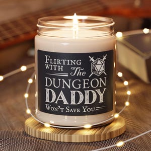 May include: A lit candle in a glass jar with a black label. The label reads "Flirting with The Dungeon Daddy Won't Save You" in white text, with a D20 die and crossed swords graphic. The candle sits on a wooden coaster, with string lights around it.