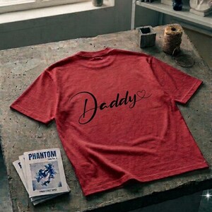 May include: A red short-sleeved t-shirt featuring the word Daddy in black cursive script with a small heart icon at the end. The shirt is made of a textured fabric and is displayed flat, showcasing the graphic print clearly on the back surface.