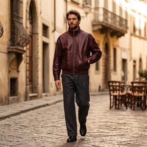 May include: A dark burgundy leather jacket with a high collar and a central zipper closure, paired with charcoal gray trousers and black leather shoes. The jacket features long sleeves with ribbed cuffs and a smooth, polished finish throughout.
