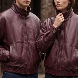 May include: Two matching deep burgundy leather jackets featuring a high stand-up collar, a full-length front zipper, and two side pockets. The jackets have long sleeves with buttoned cuffs and a ribbed elastic hem at the waist for a structured fit.