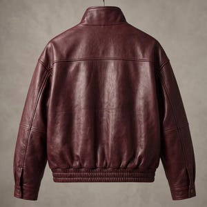 May include: A deep burgundy leather jacket featuring a stand-up collar, long sleeves with buttoned cuffs, and a ribbed elasticized hem. The back panel displays horizontal seam detailing across the shoulders and a smooth, consistent finish throughout.
