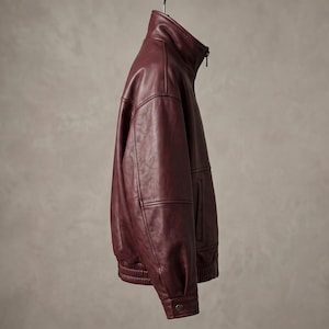 May include: A deep burgundy leather jacket featuring a high stand-up collar, a full-length front zipper, long sleeves with buttoned cuffs, and a ribbed elastic hem. The jacket is captured from a side profile view, showcasing its smooth texture.