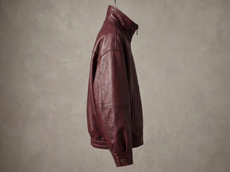 May include: A deep burgundy leather jacket featuring a high stand-up collar, a full-length front zipper, long sleeves with buttoned cuffs, and a ribbed elastic hem. The jacket is captured from a side profile view, showcasing its smooth texture.