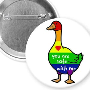 May include: A white button pin with a cartoon goose in rainbow colors and the text "you are safe with me." The goose has a red head, yellow neck, and a green body. The pin has a metal back with a safety pin.