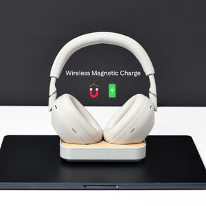 May include: Off-white wireless headphones on a charging base, resting on a black surface. The text "Wireless Magnetic Charge" is above the headphones, with a magnet and battery icon. The headphones are a modern design.