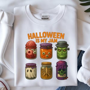 May include: White sweatshirt with the text "HALLOWEEN IS MY JAM" and illustrations of spooky jars. The jars contain a brain, eyeballs, a green monster, a ghost, a skeleton, and a skull. A pair of white sneakers and blue jeans are visible.