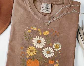 May include: A brown Comfort Colors t-shirt featuring a floral design with daisies, yellow flowers, and pumpkins. A silver necklace with a pendant is draped across the shirt. A brown hat and dried leaves are in the background.
