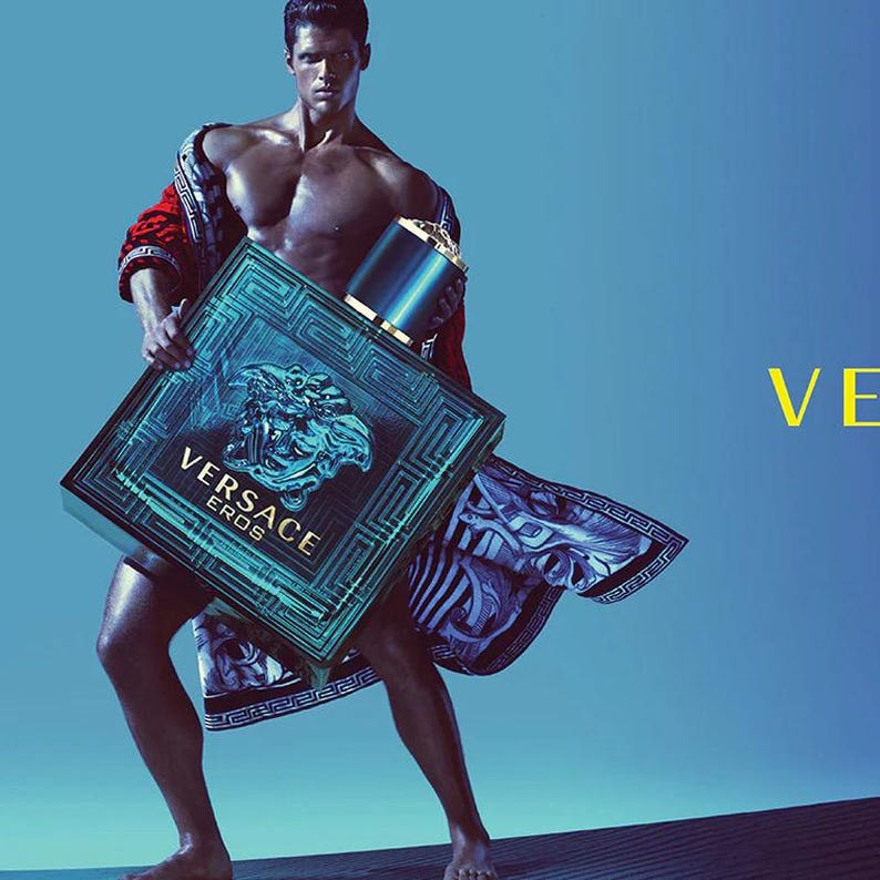 Puede incluir: Una botella y caja de perfume Versace Eros turquesa y negro son sostenidas por una persona. La caja presenta el logotipo de Versace y la palabra "EROS". La persona lleva una bata estampada en rojo y negro. El fondo es un degradado de azul.