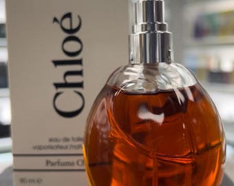 May include: A Chloé eau de toilette perfume bottle with a silver atomizer and a light brown liquid. The bottle is next to a white box with the Chloé logo in black. The bottle is 90 ml.
