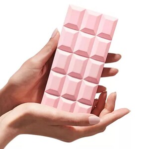 Puede incluir: Un objeto rectangular rosa con un patr&oacute;n de cuadr&iacute;cula, que se asemeja a una tableta de chocolate, se sostiene en dos manos. El objeto tiene un acabado mate y es de color rosa uniforme. Las manos son de piel clara y el fondo es blanco.
